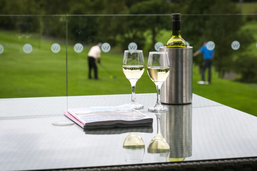 Magazines, wine glasses and wine bottle in an ice bucket sitting on an outdoor glass table overlooking a golf course